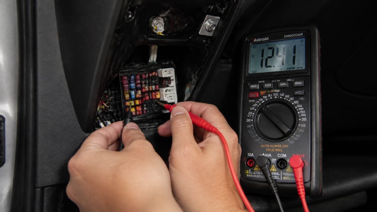 A high-resolution close-up of a technician's hands using a professional digital multimeter to test a vehicle's electrical circuit during dash cam installation, highlighting precision wiring and electronics expertise.